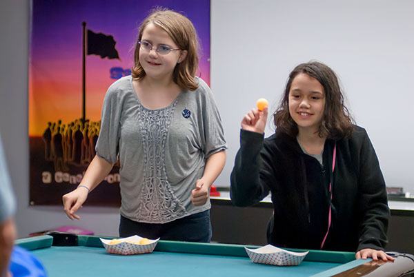 Alaura Hall and Lynzie Kenny race each other to see who can bounce the most ping pong balls across a table and into plastic buckets held by adults on the other side.